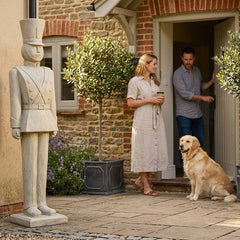 Couple with a dog standing outside a house with a garden statue and plants.