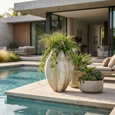 Three large decorative planters arranged beside a swimming pool on a patio, including a tall textured cream vase with ornamental grass and trailing greenery, a matching textured planter with ferns, and a round concrete planter with succulents.