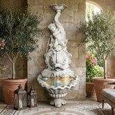 Decorative fountain with statues in a stone-walled room with plants and a sofa.