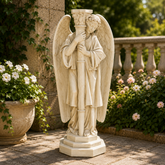 Statue of an angel holding a column in a garden setting with flowers and plants.