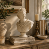 A classical-style stone bust sculpture displayed on a stack of books on a wooden console table, surrounded by home decor including a vase with greenery, a candle holder, framed art, and a bowl.