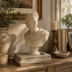 A classical-style stone bust sculpture displayed on a stack of books on a wooden console table, surrounded by home decor including a vase with greenery, a candle holder, framed art, and a bowl.