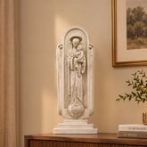A white carved religious statue of the Virgin Mary holding the infant Jesus stands on a wooden console table in a styled living room. The scene includes a beige sofa with a floral pillow, potted plants, framed wall art, and a table lamp.