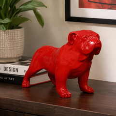 Red bulldog figurine on a wooden surface with books and a plant in the background
