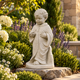A light gray stone garden statue of a kneeling child praying, displayed outdoors on a stone base in a beautifully landscaped garden with white hydrangeas, purple flowers, and green shrubs.