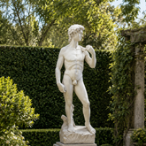 A classical-style nude male statue on a stone pedestal, displayed outdoors in a landscaped garden with trimmed hedges, potted plants, and stone paving.