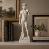 A white marble-style statue of Michelangelo's David stands on a mantel in a styled interior setting, surrounded by books, framed art, a small plant in a ribbed vase, and a lit candle against a warm gray wall.
