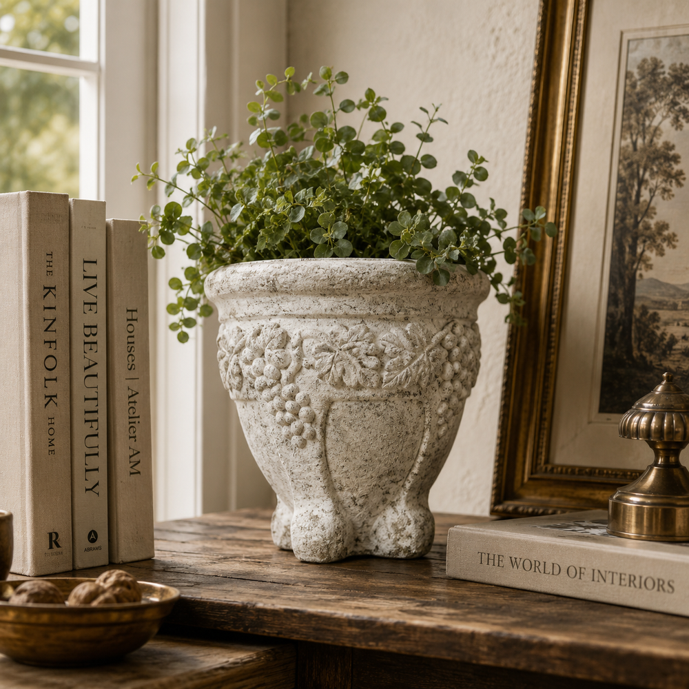 A textured stone planter with embossed grapevine details, filled with green leafy stems, styled on a rustic wooden table next to books and a framed picture.