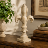 Decorative stone Fleur-de-Lys sculpture on a wooden surface with books and a plant in the background.