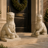 A pair of large white stone lion statues sit on either side of a dark front door, decorated with a green wreath and small lit Christmas trees.