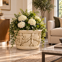 A large beige stone planter with carved cherub figures, filled with white hydrangea flowers and green foliage, styled in a bright, modern living room next to a sofa.