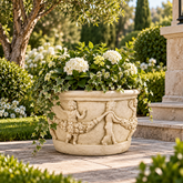 An ornate beige stone planter with carved cherub and grapevine details, filled with white flowers and green ivy, set in a lush garden next to a stone pillar.