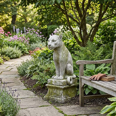 Garden scene with a stone cat statue and wooden bench surrounded by plants and flowers.