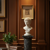 A classical white marble bust sculpture displayed on a dark marble pedestal in an elegant room with dark wood paneled walls, framed art, a round wooden table with books, and a potted plant.