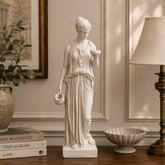 Statue of a classical woman holding a bowl on a wooden surface with books and decor items.