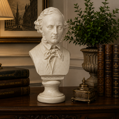 A white marble-style bust sculpture of Mende Edouard on a pedestal, displayed on a dark wooden console table in an elegant room setting with a lamp, books, and a potted plant.