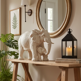 A cream-colored elephant and baby elephant statue displayed on a wooden console table in a styled room setting with a mirror, wall sconce, framed fern print, and a black lantern candle holder.