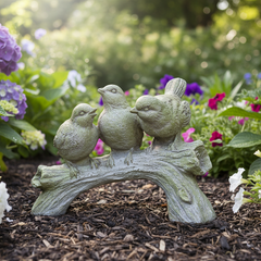 Statue of three birds on a branch in a garden setting with flowers and mulch.