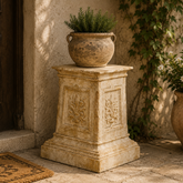 Decorative stone pedestal with a pot of plants against a rustic wall.