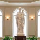 Statue of a man holding a child on a wooden pedestal in a room with arched windows and potted plants.