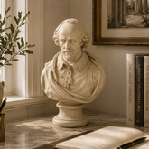 A cream-colored marble bust sculpture of William Shakespeare displayed on a marble desk in a sunlit room, surrounded by books, a potted plant, and an open notebook.