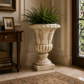 An ornate cream-colored stone-look urn planter filled with green ferns, placed in a classic entryway next to a dark wood console table and near a doorway.