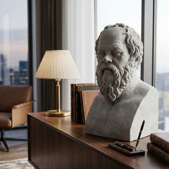 Bust of a classical philosopher on a wooden desk with books and a lamp in a room with large windows.