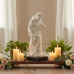 A white religious statue of Saint Christopher carrying a dog, displayed on a wooden stand with lit candles, potted plants, and roses in a softly lit interior setting.