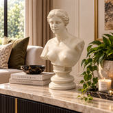 A white classical-style female bust sculpture displayed on a marble-topped console table in a styled living room setting, with books, a bowl, a potted plant, and a candle nearby.