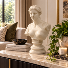 A white classical-style female bust sculpture displayed on a marble-topped console table in a styled living room setting, with books, a bowl, a potted plant, and a candle nearby.