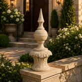 A decorative stone garden finial with a pointed top and ornate carved base, displayed on a stone wall in a landscaped garden setting with flowering bushes and a house entrance in the background.