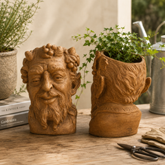 Terracotta planters shaped like faces on a wooden surface with books and scissors.