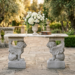 A classical stone console table with lion and winged figure supports, styled with a large white floral arrangement and a small book in an outdoor garden courtyard.