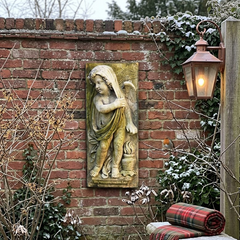 Decorative garden scene with angel sculpture on a brick wall, stone bench, and lantern.