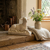 Decorative scene with a stone sculpture of a sheep, a vase of flowers, and a blanket on a wooden surface.