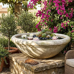 A large round stone planter bowl filled with water and flowers, placed on a stone pedestal in a sunny garden patio setting with a wicker armchair, potted plants, and bright pink bougainvillea flowers.