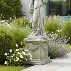 A classical-style stone statue of a draped female figure on a decorative pedestal, placed in a landscaped garden with green shrubs, lavender, white flowers, and a paved walkway.