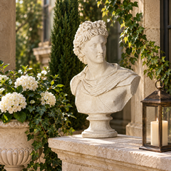 A classical-style stone bust statue of a young man with curly hair, displayed on a stone ledge in an outdoor garden setting with white flowers, greenery, and a lantern candle.