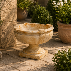 A round stone pedestal planter with a flared rim, styled outdoors on a stone patio next to potted plants and a textured stone wall.