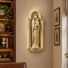 A cream-colored stone or plaster religious wall sculpture of the Virgin Mary holding the infant Jesus, mounted on a beige wall in a warm, modern home office setting with a wooden desk, leather chair, and bookshelves.