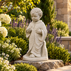A light gray stone garden statue of a kneeling child praying, displayed outdoors on a stone base in a beautifully landscaped garden with white hydrangeas, purple flowers, and green shrubs.