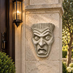 A carved stone or concrete grotesque face wall sconce mounted on a beige stone pillar outside a house, with a dark wooden door, wall lantern, and greenery in the background.