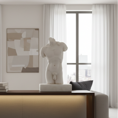 A white classical-style male torso sculpture displayed on a console table in a modern, sunlit living room with white curtains, a framed abstract painting, and a dark gray pillow.