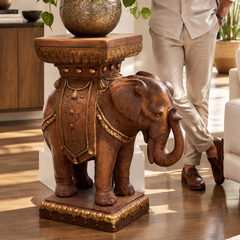 A man in a light beige lounge set stands holding a mug near a large elephant-shaped wooden side table holding a potted green plant, while a woman sits on a white sofa in a bright modern living room.