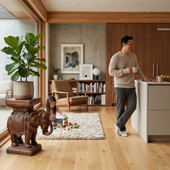 Elephant Pedestal A man in a beige sweater and gray pants stands in a modern kitchen area holding a white mug, with a young girl playing with blocks on a white rug nearby. The room features light wood floors, a concrete wall, and a large potted fiddle-leaf fig plant on a decorative elephant pedestal.