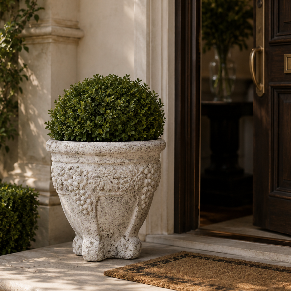 A spherical green topiary tree in a textured, carved light gray stone planter, placed on a stone doorstep next to an open dark wood front door.