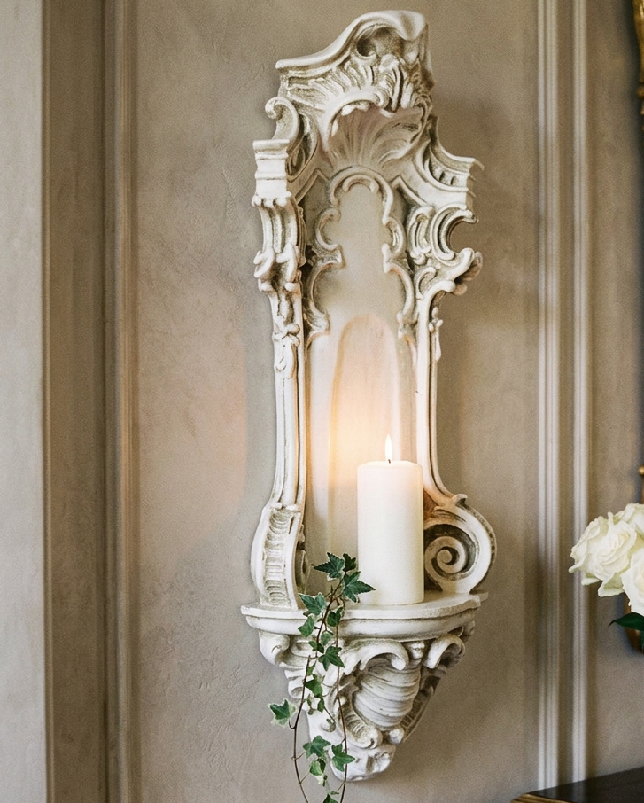 An ornate white carved wall sconce holding a lit white pillar candle, mounted on a beige wall beside a gold-framed mirror and a table with white roses.