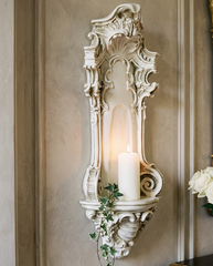 An ornate white carved wall sconce holding a lit white pillar candle, mounted on a beige wall beside a gold-framed mirror and a table with white roses.