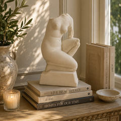 A cream-colored stone sculpture of a seated nude female figure displayed on a stack of books on a wooden table, next to a lit candle, a potted plant, and a small bowl in a sunlit room.