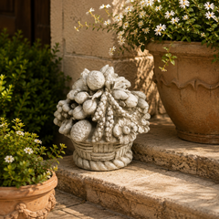 A decorative stone sculpture of a basket overflowing with grapes and round fruits sits on outdoor stone steps beside potted plants with white flowers.